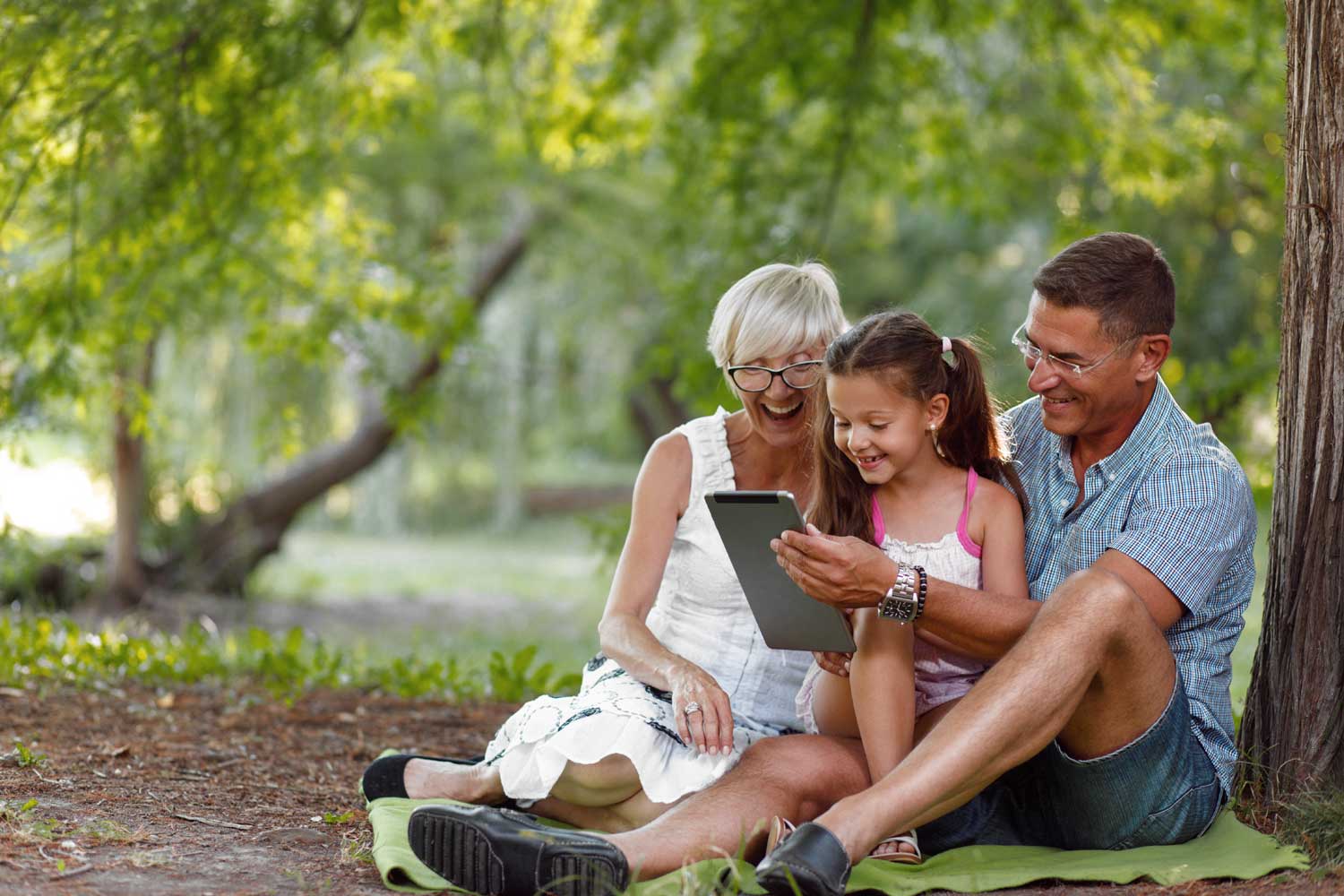 Großeltern mit Enkeltochter in der Natur unter Bäumen sitzend freudig auf ein Tablet sehend